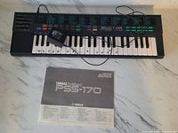 Top-down view of Yamaha PSS-170 keyboard connected with power cord, original manual laid out below on marble surface