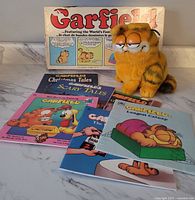 Photo of 9-inch Garfield plush, board game box, and six Garfield softcover books showing front covers and overall condition