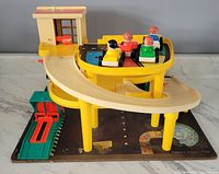 Front angled view showing yellow parking garage structure with attached beige ramp, green elevator, and four Little People figures in cars on the top parking level.