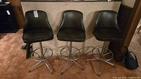 Three dark brown leather-look cushioned swivel bar stools with chrome metal legs and footrests shown together from front.