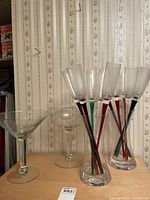Photo displaying a group of stemmed champagne flutes with colored stems inside two clear glass vases beside a large martini glass and a large champagne glass on a wooden surface against floral wallpaper.