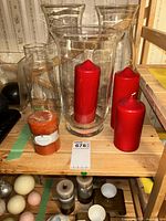 Photo of three clear glass hurricane candle holders and four candles (three red, one orange) arranged on a wooden surface with a numbered tag in front.