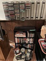 Wall-mounted CD racks partially filled with clear plastic jewel-cased CDs above a woodgrain panel wall, shelf below with DVDs and loose media on floor
