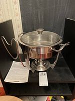 Front angled view of the Tramontina chafing dish showing the stainless steel frame, glass lid with knob, and lid holder