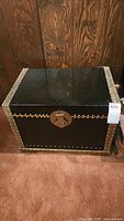 Full view of the black lacquered chest with metal rivets on corners and edges, sitting on carpeted floor