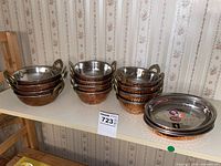 Photo showing 24 copper bowls on a shelf: 12 with double brass-colored handles and 12 without handles stacked in groups. Bowls have hammered copper exteriors and smooth stainless steel interiors.