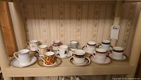 Shelf holding 12 assorted demitasse teacups and saucers from various brands including Royal Crown Derby, Wedgewood, Paragon, Royal Doulton.