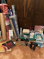 Photo showing assorted rolls of gift wrapping paper, glue guns, scissors, ribbons, crafting books and other crafting tools and materials arranged on a wooden surface.