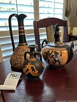 Three Grecian-style ceramic pieces arranged on a wooden table by a window: a tall jug, a small vase, and a covered jar