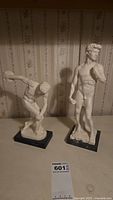 Two mini Greek statues on black bases: one discus thrower in mid-throw and one standing male figure.