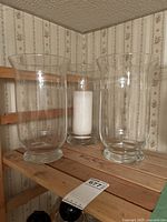 Three clear glass hurricane candle holders on a wooden shelf; one contains a white pillar candle