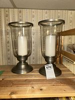 Front view of pair of hurricane candle holders with white pillar candles on wooden surface.