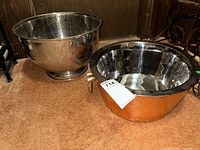 Two ice buckets displayed side by side on brown carpet: hammered copper double-walled ice bucket with handles and a large silver-tone bucket without handles.