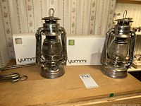 Pair of Dietz lanterns with glass globes on wooden surface, two metal hangers coiled beside them, background shows boxes of Yummi candles