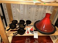 Le Creuset Moroccan tagine with red enamel conical lid, black base, seven mini cast iron frying pans and a cooking guide on a wooden shelf.