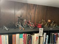 Seven miniature decorative bicycles displayed on a shelf above books, various styles and colors