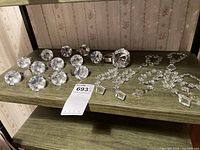 Overhead view showing 12 small napkin rings with large clear faceted crystals and 1 larger napkin ring alongside 7 chandelier-style glass place card holders on a wooden shelf.
