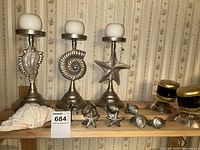 Front view of three silver-colored metal candle holders with ocean-themed designs, each topped with a round white candle, alongside six matching ocean-themed metal napkin rings and a single large seashell.