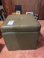 Front view of green/grey upholstered storage ottoman with cushioned top showing four stitched sections.