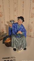 Front-left angled view of Royal Doulton figurine depicting elderly lady with birds and basket of produce, displaying detailed colors and craftsmanship.