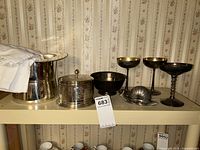 Silverplate top hat ice bucket, covered snail condiment dish, goblets, and bowl arranged on a shelf against wallpaper backdrop.