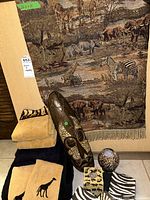 Photo of the entire set displaying the tapestry hung on the wall, African mask placed in front along with neatly folded hand towels in beige, black, and tiger print. Glass vase and animal print candle holder alongside zebra print napkins arranged on floor.