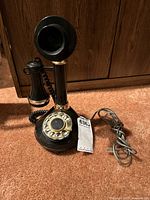 Black vintage style candlestick telephone with rotary dial laid on orange carpet, showing front and side views with attached cords.