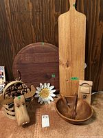 Photo showing long rectangular Jamie's Italian charcuterie board leaning upright, large round wooden board behind it, wooden salad bowl with utensils, carved decorative moose head, wooden flower, and small wooden spice mill grouped together on floor.