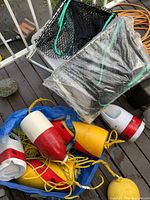 Blue bag containing buoy floats in red, white, and yellow with attached ropes, prawn traps with metal frames and black nets, and plastic-wrapped bag with sardine cans.