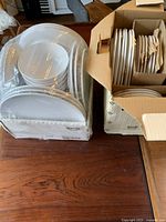 Two cardboard IKEA boxes containing white ceramic dinnerware sets, wrapped in clear plastic. One box shows bowls on top, the other plates organized vertically.