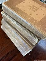 Four hardcover antique dictionaries stacked, showing beige patterned cloth covers with wear and fraying on edges.