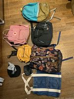 Lot of backpacks and bags arranged on floor, showing five backpacks, helmet bag, tote bag, and cap.