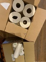Box of 6 large industrial paper towel rolls, white with cardboard cores, arranged inside an open cardboard box on a floor surface.