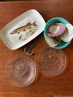 Full view of lot items on wooden surface showing ceramic fish plate, glass crab plates, seashells in a green bowl, and metal anchor hook.