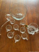 Full view of punch bowl and eight matching glass cups with clear handles, one cup cracked, arranged on a wooden surface.