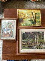 Three framed art pieces on wooden table: two landscape oil paintings and one print of a man asleep at desk