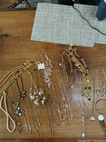 First photo shows a large assortment of necklaces on a wooden table, featuring gold-tone chains, faux pearls, and bead designs.