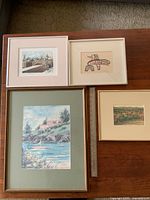 Four framed artworks on wood table showing a coastal watercolour, an indigenous style silkscreen, and two other framed prints including one small landscape.