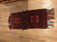 Pair of rectangular Baluch Khorjin pouches laid out flat on wooden floor, displaying deep red and black geometric patterns, connected by loops of camel hair on the center seam, fringed ends with tassels.