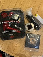 Photo showing the 6-piece wine tool kit in its clear plastic case alongside the black UV HVAC sterilization device and accessories on a wooden floor.