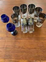 Photo showing 8 Dorothy Thorpe clear glass tumblers with silver floral/damask pattern and 2 cobalt blue stemmed wine glasses on wooden table.