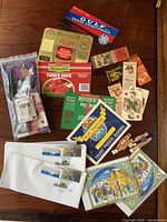 Full lot overview showing first day covers, food labels, matchbook covers, playing cards, and a packaged souvenir item on wooden surface.