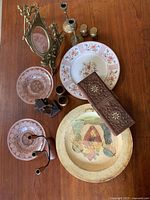 Overview showing most items including decorative plates, pink glass plates, wooden carved box, brass candle holder, and brass salt and pepper shakers