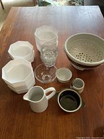 All items displayed on wooden table, includes 8 white Arcopal milk glass bowls, clear glass bowl, small white ceramic cups, pitcher, brown glazed bowl, and ceramic colander.