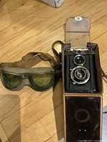 Shows the US Army vintage goggles with green lenses and Voigtländer camera in leather case on wooden floor.