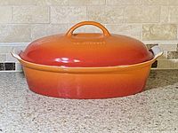 Le Creuset oval casserole dish with matching lid, gradient orange to red exterior and cream interior, placed on kitchen countertop.