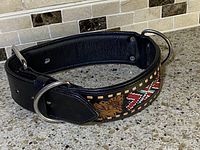 Full view of large black leather dog collar with beadwork and metal rings lying flat on a stone surface.