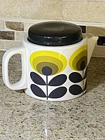 Front side of the Orla Kiely teapot showing yellow and olive green floral design with black petals and round black lid on white ceramic body.