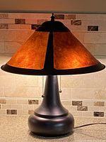 Front view of MCM style lamp showing conical brown Mika style shade with black metal strips and black rounded metal base on granite countertop with tile backsplash.