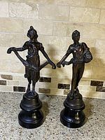 Pair of metal antique French spelter spelter figures of a female and male with black wooden bases, front angle view on tiled backsplash.